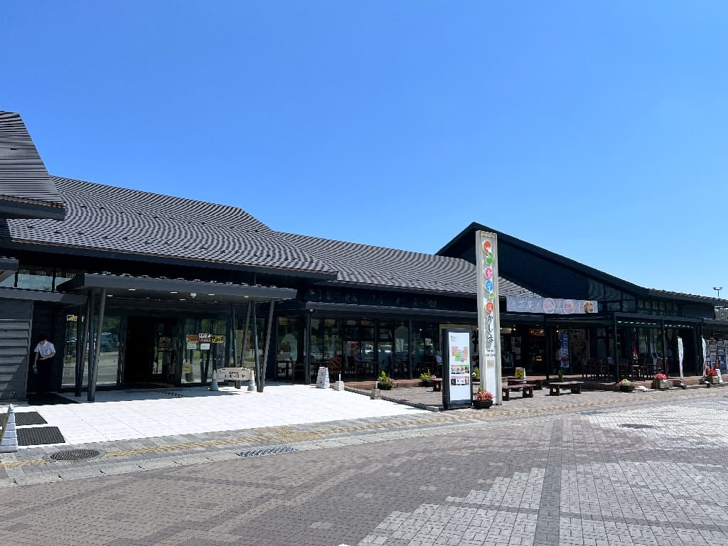Ultra-realistic high-quality photograph of the Minamisoma-Kashima Service Area under a pristine blue sky. The image features a traditional yet modern Japanese-style building with a dark tiled roof, accompanied by a spacious, bright stone-paved walkway. 抜けるような青空の下に佇む、常磐自動車道・南相馬鹿島SA（セデッテかしま）の全景。黒い瓦屋根を戴く和風モダンな平屋建ての建築と、手前に広がる明るい石畳のコントラストが美しい情景