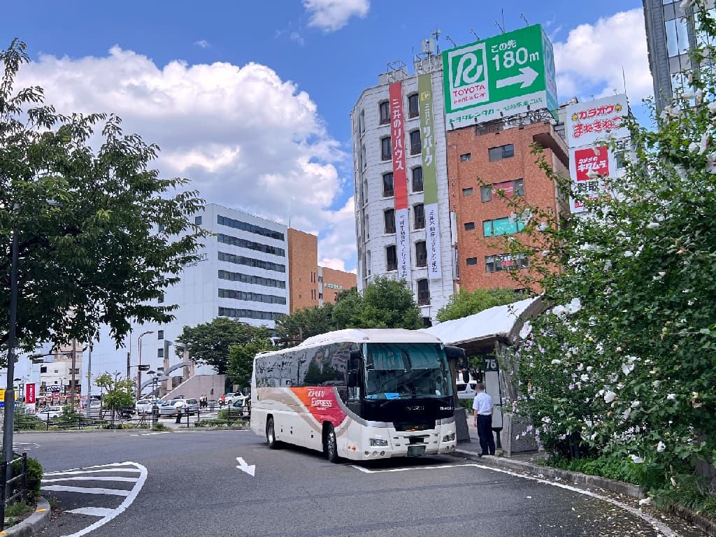 Ultra-realistic high-quality photograph of a white Tohoku Express bus with red and orange stripes parked at the Sendai Station East Exit terminal under a clear blue sky. A driver is standing nearby, courteously seeing off the arriving passengers as they alight, capturing the authentic atmosphere of the journey's end. 晴天の仙台駅東口バスターミナルに到着し、停車している白地に赤とオレンジのラインが入った東北急行バスの車両。手前には乗務員が立っており、降車されるお客様へ感謝を込めて丁寧にお見送りをしている臨場感が伝わる情景
