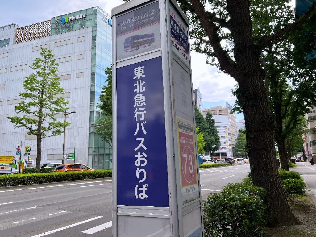 Ultra-realistic high-quality photograph of the Tohoku Express Bus stop sign at Hirose-dori 41 in Sendai, prominently displayed in blue and white. The background features lush street trees and the MetLife building, clearly depicting the surrounding urban environment. 仙台市内の広瀬通りに立つ「東北急行バス 広瀬通り41番おりば」の案内板。青地に白文字で視認性が高く、背景には緑豊かな街路樹とメットライフ仙台ビルが写り込んでおり、実際の乗車場所の周辺環境が明確に伝わる情景