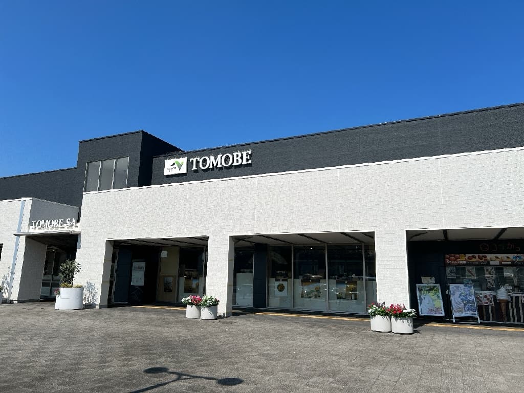 Ultra-realistic high-quality photograph of the modern and chic exterior of the Tomobe Service Area on the Joban Expressway. The black and white building design is beautifully accented by colorful flowers in planters near the entrance. 常磐自動車道・友部SA（TOMOBE SA）の洗練された外観。白と黒を基調とした現代的でシックなデザインの建物で、エントランス付近には色彩豊かな花が植えられたプランターが配置されている